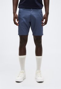 Άνδρας που φορά μπλε σορτς, λευκές αθλητικές κάλτσες και λευκά αθλητικά παπούτσια, στέκεται μπροστά σε μια απλή ανοιχτόχρωμη επιφάνεια.