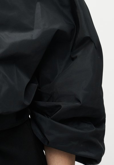 Manche bouffante noire avec plis et rides visibles sur un vêtement, couvrant partiellement le bras sur un fond clair uni.