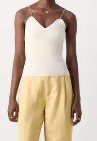 Ribbed cream tank top with a V-neck and chain strap accents, paired with loose-fitting, light yellow pants. Smooth texture throughout.