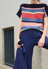 Persoon in een multicolore gestreepte trui met korte mouwen en een marineblauwe broek, houdt een marineblauwe blazer vast, staat naast een stenen muur met een beglaasde vensterbank.