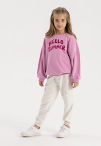 Rosa Sweatshirt mit "HELLO SUMMER" in fetten Buchstaben, kombiniert mit weißen Jogginghosen und weißen Sneakers. Glatter Stoff, lässige Passform.