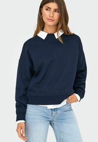 Felpa oversize blu navy con colletto bianco, polsini e orlo a coste, abbinata a jeans azzurri. Tessuto con trama liscia.