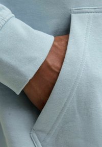 Matière de sweat-shirt bleu clair avec une texture lisse, mettant en avant un gros plan d'une main glissée dans une poche avant, détails de couture visibles.