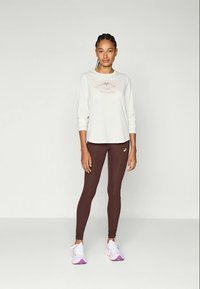 Pitkähihaisen kermaisen urheilupaidan logo on ruskeiden leggingsejen yllä. Mallilla on päällään vaaleat lenkkarit, joissa on vaaleanpunaisia yksityiskohtia. Nahkan sileä kangas.