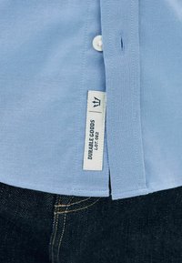 Šviesiai mėlyna marškinė su mygtukais, pagaminta iš tekstūruotos medžiagos, su etiketė, pažymėta "DURABLE GOODS LOT 082." Derinama su tamsiais džinsais.