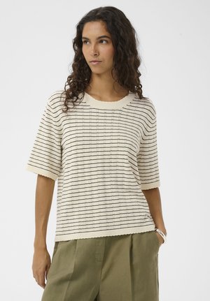 Femme aux longs cheveux bouclés portant un haut en maille à manches courtes crème avec des rayures horizontales noires et un pantalon taille haute vert olive.