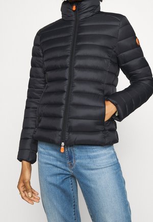 Schwarze gesteppte Jacke mit orangefarbenen Reißverschlussanhängern, getragen von einer Person in blauen Jeans, eine Hand in der Tasche, vor einem einfarbigen Hintergrund.