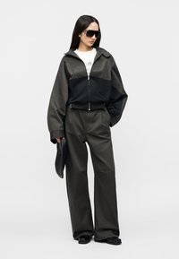 Γυναίκα με υπερμεγέθη μαύρο και σκοτεινό γκρι μπουφάν, παντελόνι με φαρδιά μπάτζα, μεγάλα γυαλιά ηλίου, κρατώντας μαύρο φάκελο, στέκεται σε απλό φωτεινό φόντο.