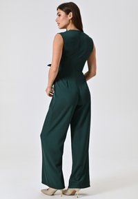 Groene jumpsuit met een aansluitend lijfje, mouwloos ontwerp, wijde pijpen en een rits aan de achterkant. Draagt hij met lichtgekleurde hoge hakken.