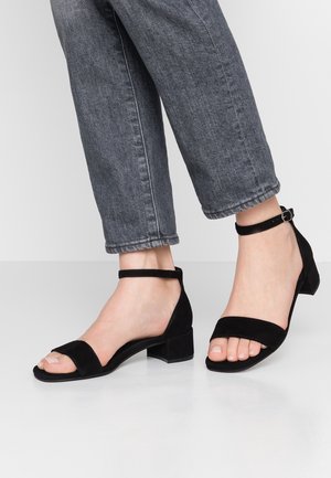 Czarne sandały na obcasie zamszowe, z otwartym noskiem, paskiem wokół kostki i obcasem typu blok. W połączeniu z szarymi, przykrótkimi dżinsami.