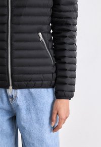 Melnas polsterētas jakas ar vertikālu quilted rakstu, ar sudraba rāvējslēdzēju un sānu kabatu, apvienota ar gaiši ziliem džinsu biksēm.