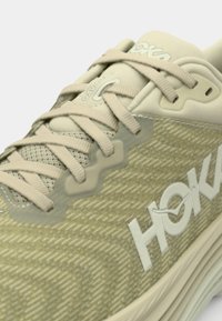 Beige løpesko med teksturert overdel, mesh-detaljer og tonede snører. Har en fremtredende hvit logo og polstret såle.