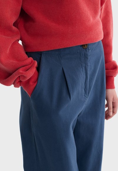 Osoba ubrana w luźną czerwoną bluzkę z długim rękawem wpuszczoną do wysokowaistowanych, niebieskich plisowanych spodni, z jedną ręką w kieszeni i zrelaksowaną postawą.