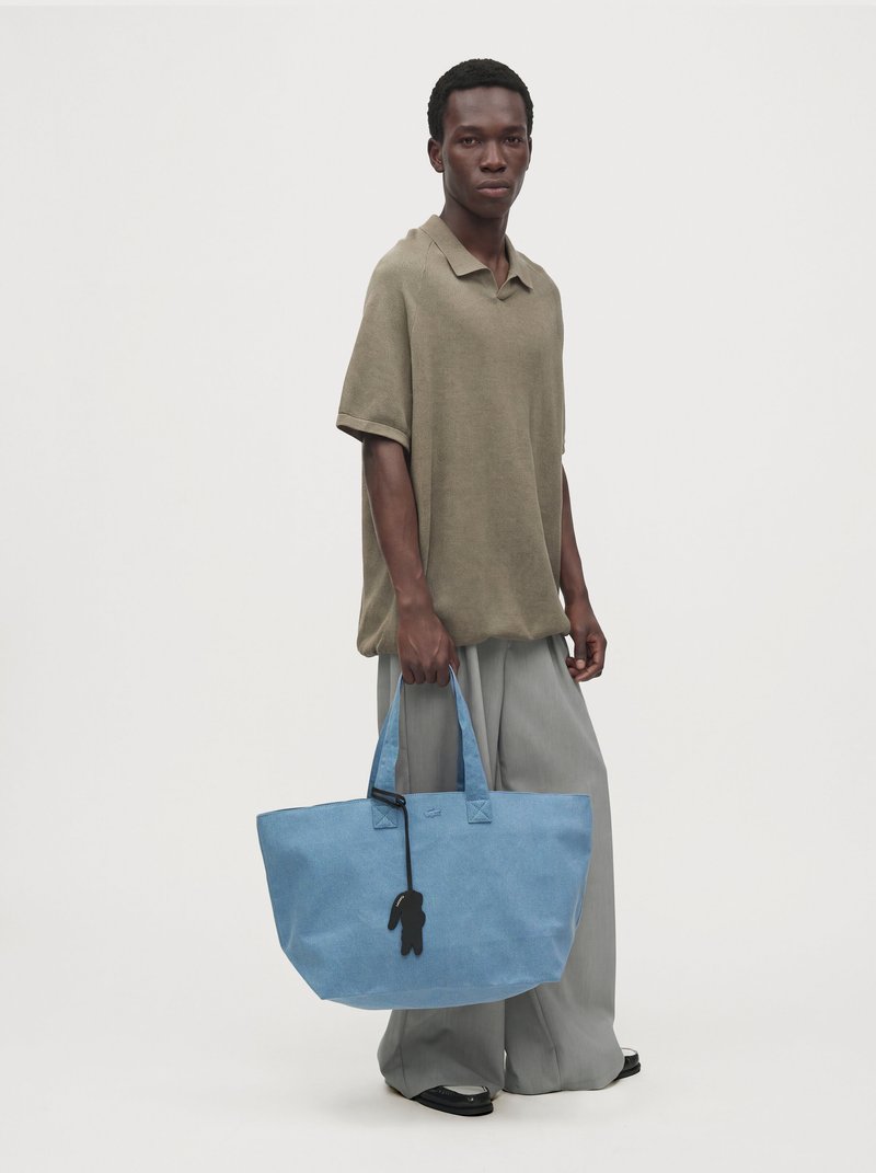 Un homme porte un polo beige ample et un pantalon gris, et tient un grand sac cabas bleu orné d’un charm noir, devant un fond de studio uni.