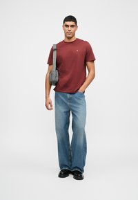 Bordová polokošeľa s krátkym rukávom, voľné džínsy a čierne topánky. Modelka nesie šedú crossbody tašku, stojí proti svetlému pozadiu.