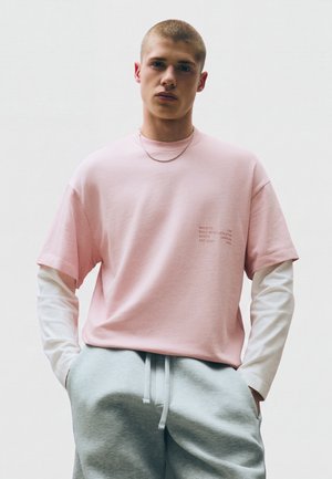 Junger Mann mit kurzen Haaren, der ein pinkes Kurzarmhemd über einem weißen Langarmshirt und grauen Jogginghosen trägt, die Hände in den Taschen.