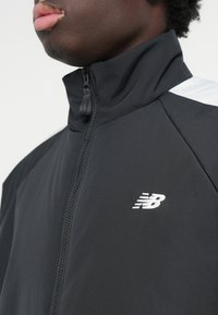 Giacca nera con zip e colletto alto, con accenti bianchi. Presenta un tessuto strutturato e un piccolo logo bianco sul petto.