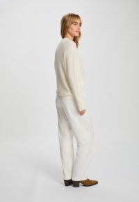 Kermainen teksturoitu neule, löysä istuvuus, yhdistetty valkoisiin leveälahkeisiin housuihin. Ruskeat nilkkurit ja rento asento, kääntyen hieman sivulle.