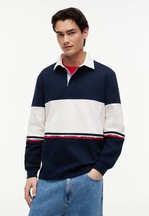 Jeune homme aux cheveux foncés portant un maillot de rugby rayé bleu marine, blanc et rouge avec un col blanc et un jean bleu clair, la main dans la poche.