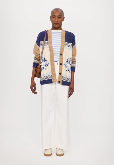 Beige und blaue Strickjacke mit floralen Mustern, Knöpfen vorne, über einem gestreiften Hemd, kombiniert mit weiten weißen Hosen und Sneakers.