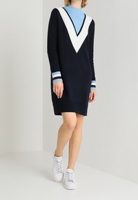 Robe tricotée marine avec un col en V blanc et des accents côtelés bleu clair au col et aux poignets. Portée avec des baskets blanches, mettant en valeur un look décontracté.