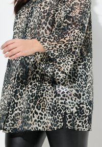 Blusa in stampa leopardata trasparente, caratterizzata da colori beige e neri, vestibilità ampia, maniche larghe e spacco laterale per una maggiore libertà di movimento.
