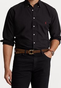 Svart skjorta med krage, uppvikta ärmar, vita knappar och röd logotyp. I kombination med brunt bälte och klocka, bärs med svarta jeans.