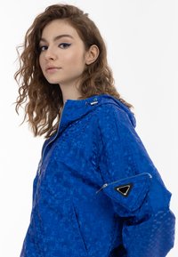 Giacca blu con cappuccio caratterizzata da un motivo geometrico, chiusura con zip, tasche laterali e una tasca con zip sul manica decorata con dettagli triangolari.