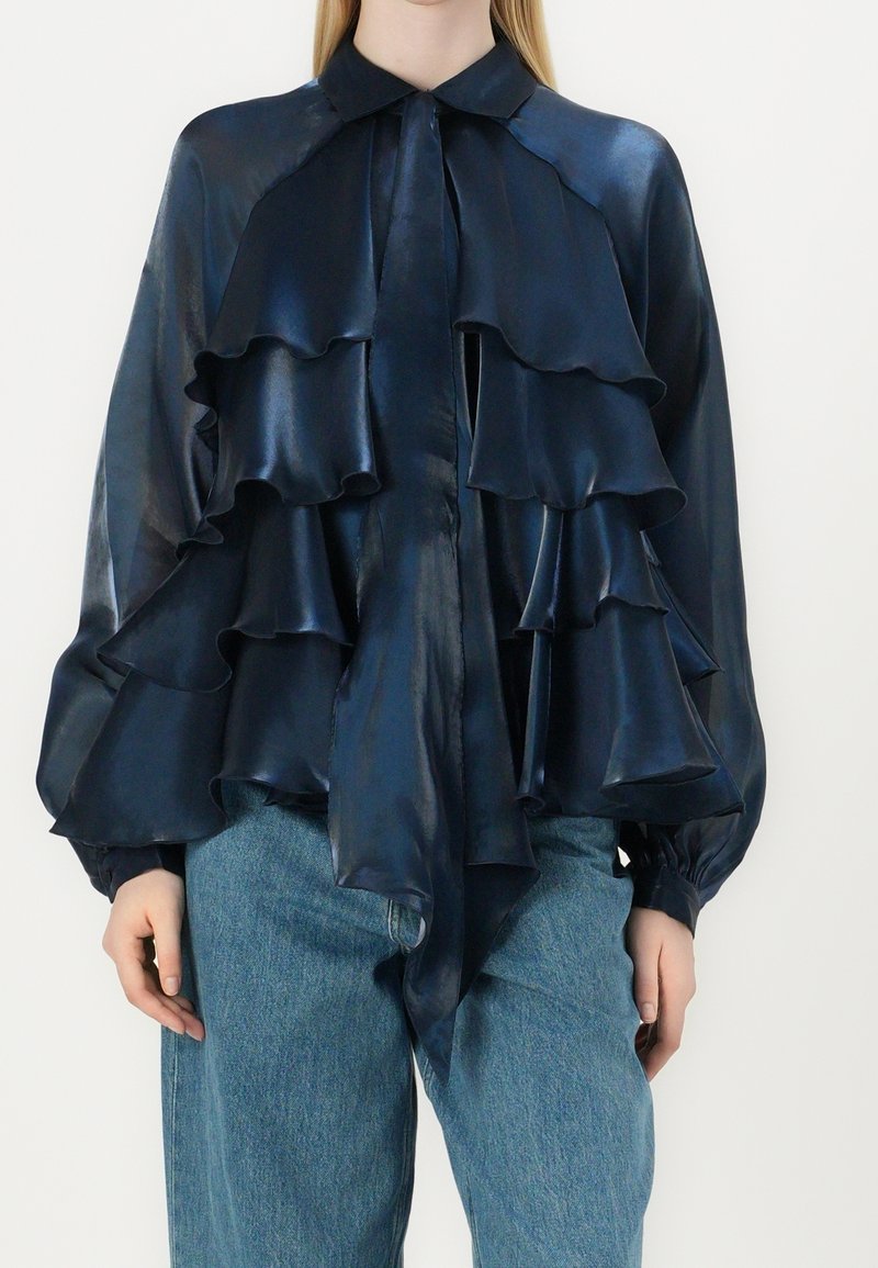 Femme portant un chemisier en satin bleu marine avec des volants superposés et des manches longues, associé à un jean en denim bleu clair.