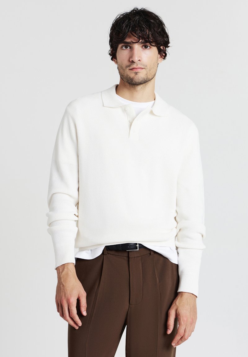 Weißes geripptes langärmliges Polohemd mit klassischem Kragen und drei Knöpfen, über einem weißen T-Shirt getragen und mit braunen Hosen kombiniert.