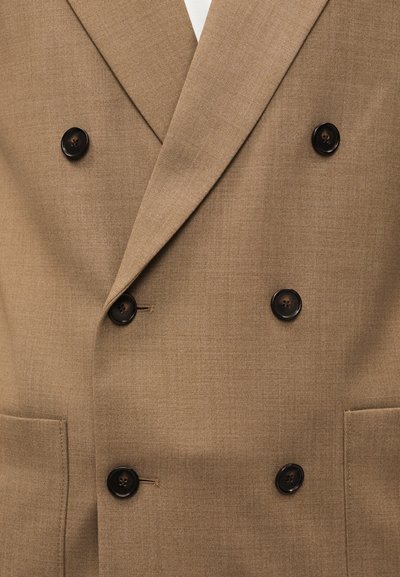 Διπλόμπροστο μπεζ σακάκι με έξι σκουρόχρωμα κουμπιά και δύο μπροστινές τσέπες πάνω σε λείο ύφασμα.