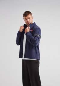 Chaqueta ligera de color azul marino con cremallera frontal, bolsillo en el brazo con cremallera y puños elásticos. Presenta un acento de logotipo rojo y un diseño de cuello alzado.