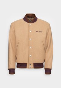 Veste en laine beige avec des boutons-pression argentés, des poignets et un ourlet côtelés violets, arborant un logo brodé "Marc O'Polo" sur la poitrine.