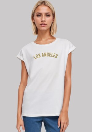 Blonde woman wearing a white sleeveless top with "LOS ANGELES" in gold text and blue jeans, standing against a plain gray background.