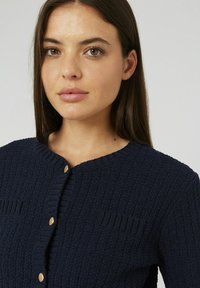 Cardigan texturé bleu marine avec un col rond, cinq boutons dorés et des détails côtelés, comprenant de petites poches à l'avant.