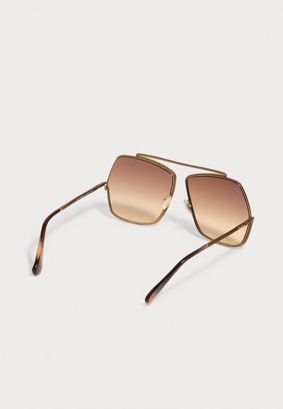 Lunettes de soleil carrées avec fines montures dorées et verres marron dégradés, posées sur une surface claire.