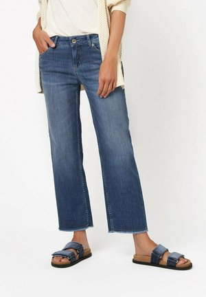 Weite Denim-Jeans in dunkelblauem Waschung mit ausgefranstem Saum, kombiniert mit marineblauen Sandalen mit gepolsterten Riemen. Das Model trägt ein kurzärmliges Oberteil.