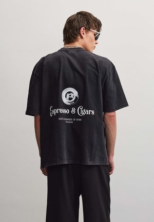 Mand iført sort oversized T-shirt med teksten "Espresso & Cigars" og logo på ryggen, kombineret med sorte bukser og solbriller, set bagfra.