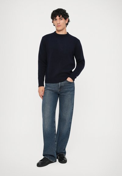 Giovane uomo con capelli ricci che indossa un maglione blu scuro, jeans a gamba larga blu e scarpe da ginnastica nere, in piedi con una mano in tasca.
