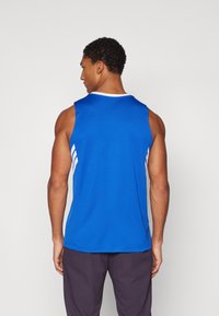 Zils sporta topiņš ar noapaļotu kakla izgriezumu, ar baltām sānu svītrām un gludu, vieglu audumu. Valkāts ar tumšiem šortiem.