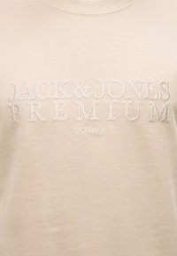 Μπεζ ύφασμα με λευκό κεντητό κείμενο «JACK & JONES PREMIUM» και μικρότερα γράμματα «EST. 1990» από κάτω.