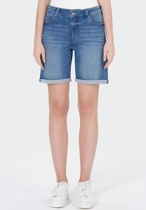 Femme portant un short en denim bleu à ourlets roulés et des baskets blanches, debout devant un fond blanc uni.