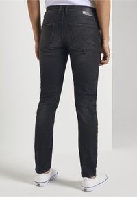 Person som bär tajta svarta jeans och vita sneakers, som står mot en ljus neutral bakgrund, sedd bakifrån.