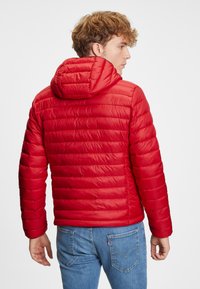 Rød puffjakke med hætte, prydet med quiltet tekstur og strømlinet design. Båret over blå denimjeans.