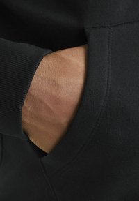 Schwarzer Hoodie-Stoff mit einer strukturierten Oberfläche; auf der Seite ist eine Tasche sichtbar, in der eine Hand ruht, die Hautton und -struktur zeigt.