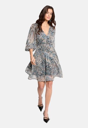 Femme portant une robe longue au genou, fluide et à motifs, avec des manches bouffantes et des talons pointus noirs, marchant avec assurance devant un fond uni.