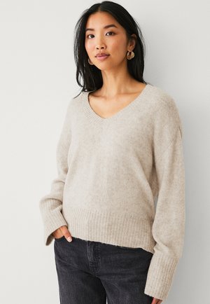 Beige gebreid sweater met een V-hals, geribbelde onderrand en lange mouwen. De stof lijkt zacht met een licht gestructureerde uitstraling. Gecombineerd met donkere jeans.