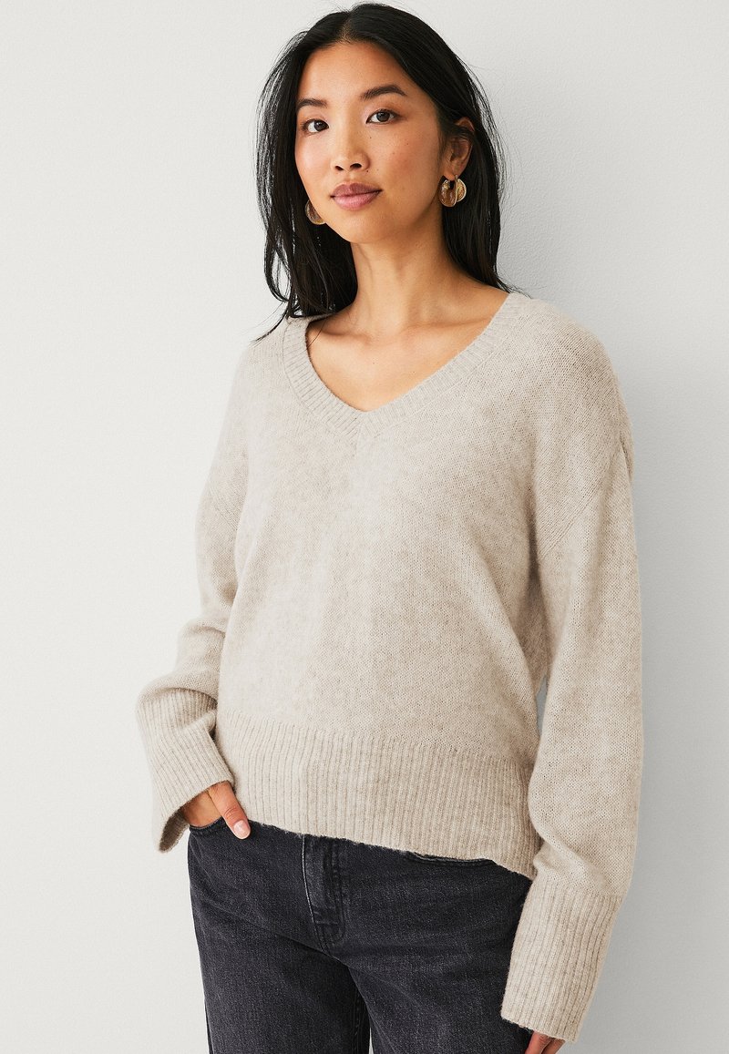 Beige gebreid sweater met een V-hals, geribbelde onderrand en lange mouwen. De stof lijkt zacht met een licht gestructureerde uitstraling. Gecombineerd met donkere jeans.