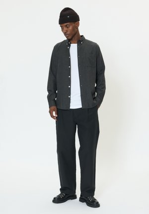 Dunkelgraues Hemd über einem weißen T-Shirt, kombiniert mit lockeren schwarzen Hosen und schwarzen Schuhen mit silbernen Akzenten, vor einem schlichten Hintergrund.