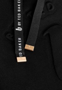 Sort stof med to sorte snore, den ene trykt med "BY TED BAKER" i hvid og metalender graveret med "BY TED BAKER" med et logo.